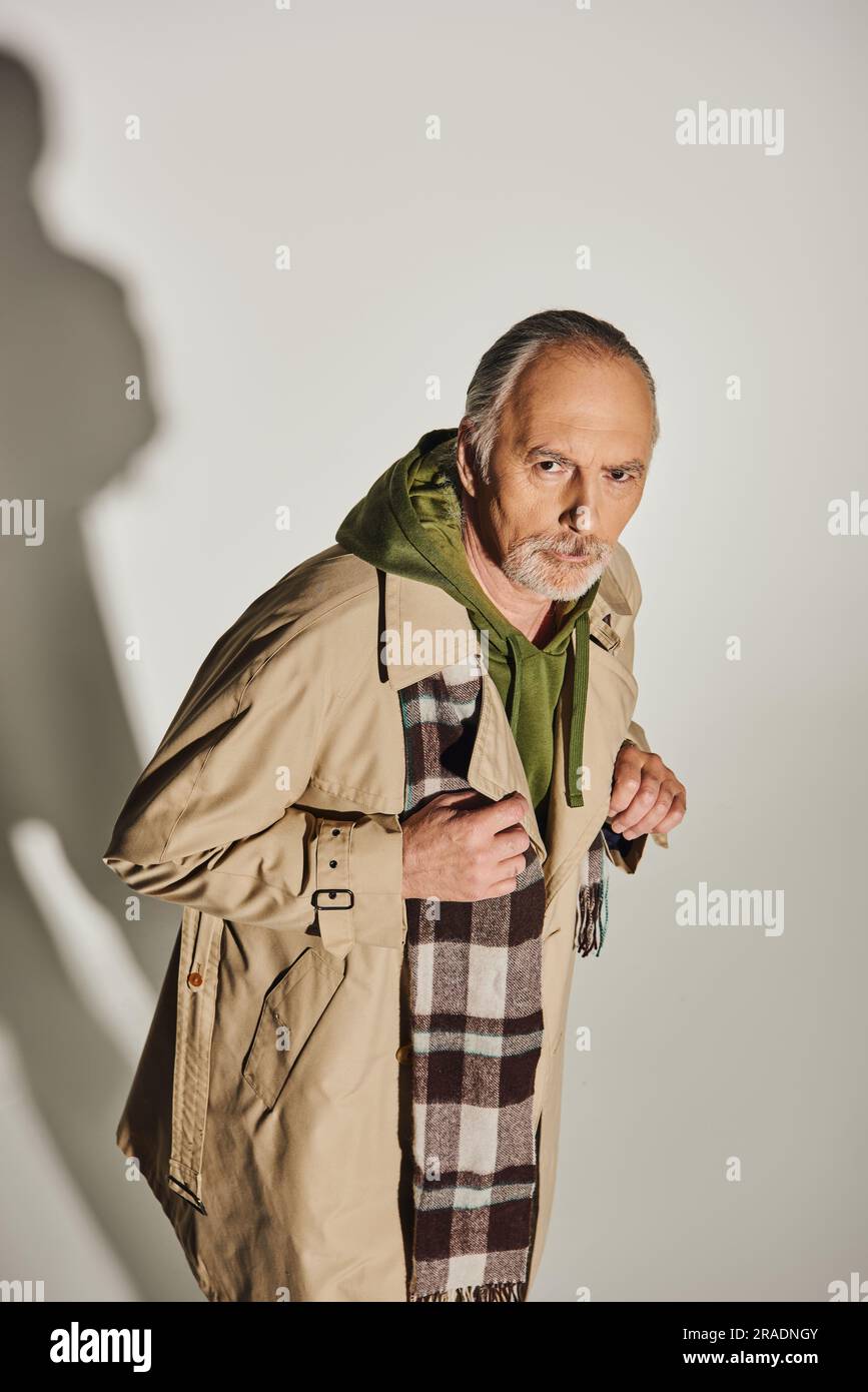 homme d'âge plein de confiance avec un regard sérieux et expressif regardant l'appareil photo sur fond gris avec ombre, vêtements décontractés élégants, pull à capuche vert, foulard à carreaux, Banque D'Images