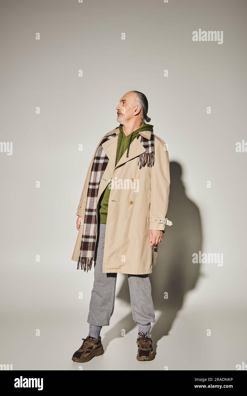 modèle homme senior en trench-coat beige, écharpe à carreaux et pull à capuche vert sur fond gris avec ombre et vue sur l'extérieur, élégant Banque D'Images