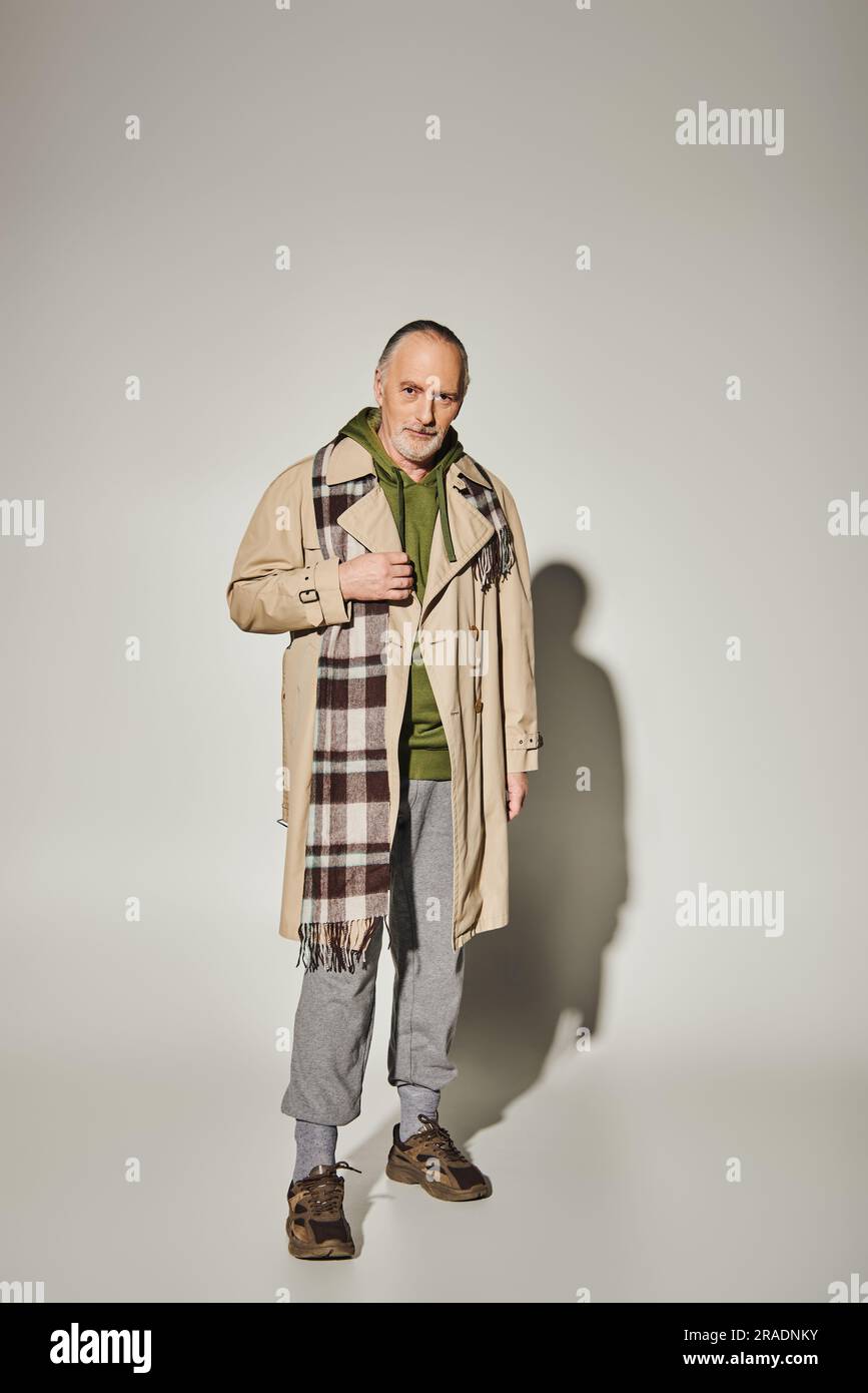 look tendance, vieillissement positif, modèle masculin senior souriant à capuche vert, trench beige et foulard à carreaux debout et regardant venu Banque D'Images