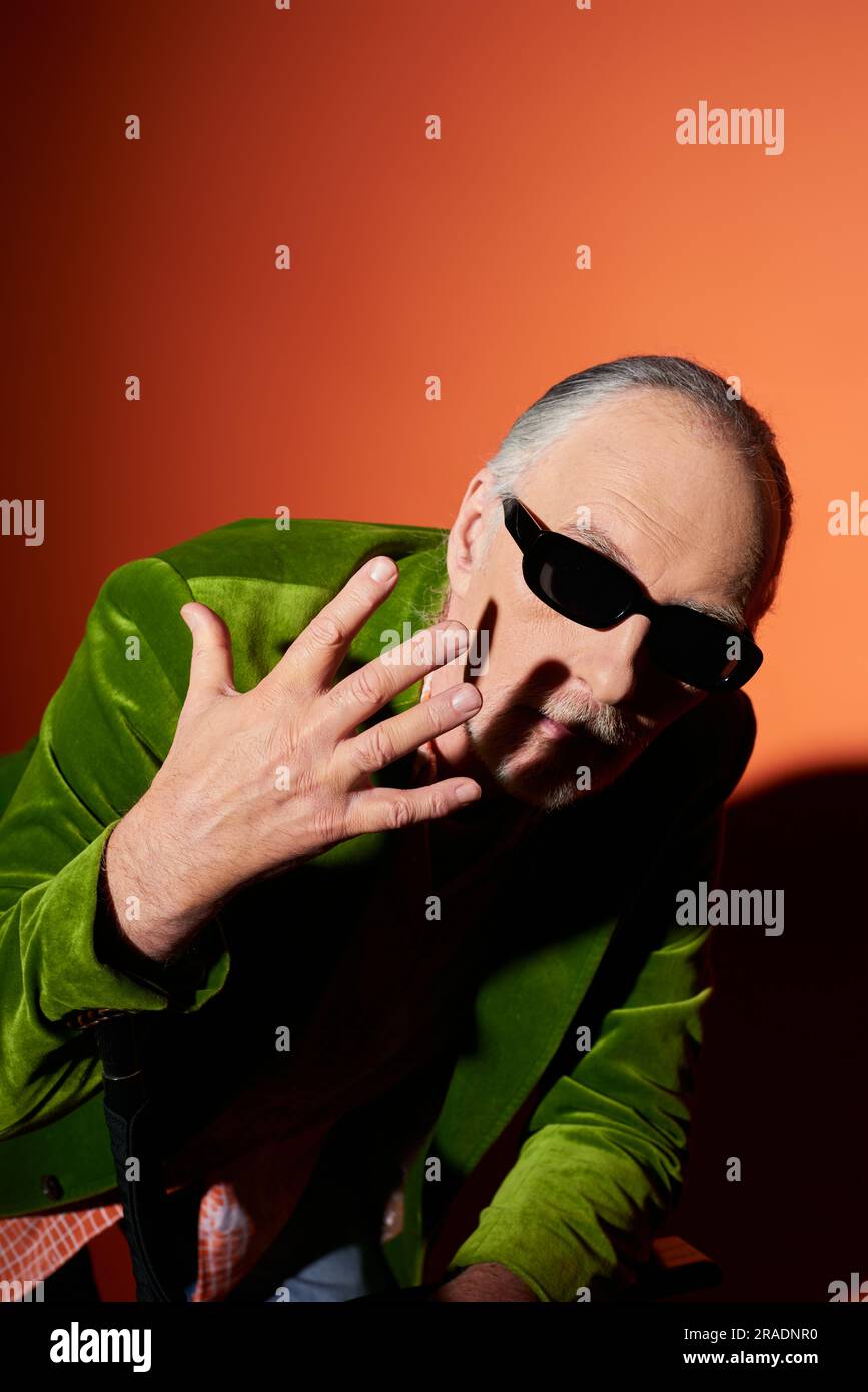 modèle masculin senior expressif en lunettes de soleil sombres et blazer en velours vert qui gestuelle avec la main près du visage et regardant l'appareil photo sur le dos rouge et orange Banque D'Images