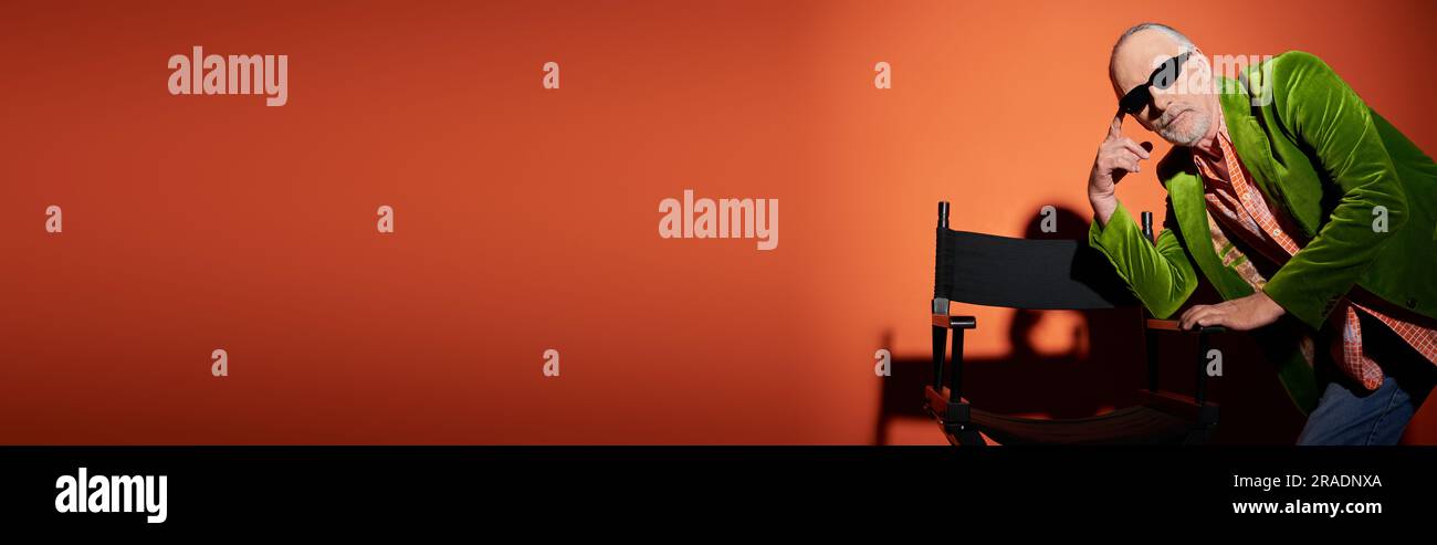 modèle masculin senior réfléchi en velours vert et lunettes de soleil foncées regardant l'appareil photo tout en posant près de la chaise sur fond rouge et orange avec sh Banque D'Images