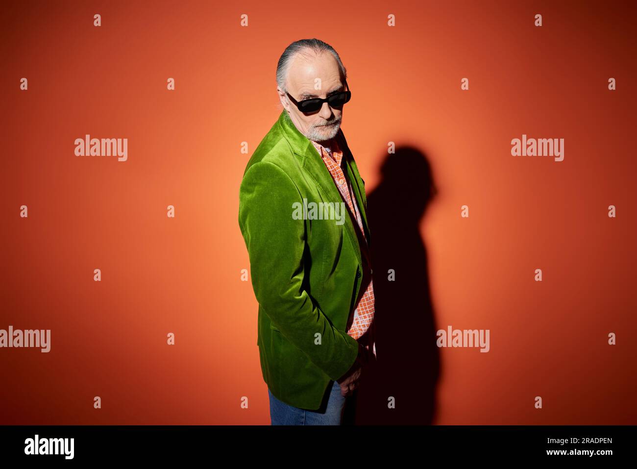 modèle senior pour homme dans des vêtements décontractés élégants regardant l'appareil photo sur fond rouge et orange avec ombre, blazer en velours vert, lunettes de soleil sombres, expressi Banque D'Images