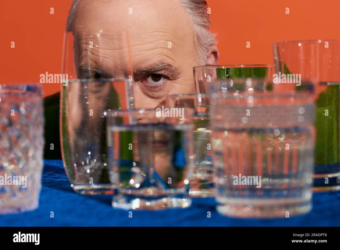 modèle masculin senior avec regard expressif regardant la caméra derrière des verres en cristal flous avec de l'eau pure sur fond orange, population vieillissante, symbole Banque D'Images