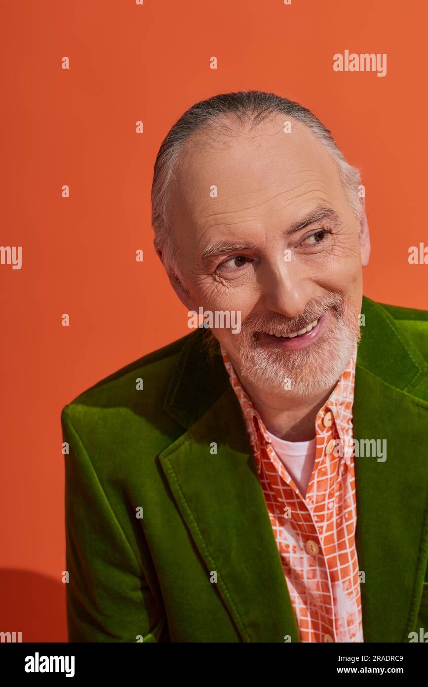 concept élégant de vieillissement et de style de vie positif, portrait d'un modèle homme senior optimiste souriant et regardant loin sur fond orange vif, gris hai Banque D'Images