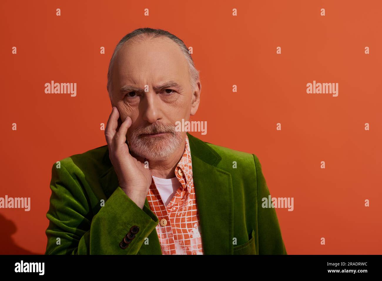 mode et âge, portrait d'un homme gris aux cheveux, barbu, réfléchi touchant le visage et regardant l'appareil photo sur fond rouge orange, modèle masculin senior, agi Banque D'Images