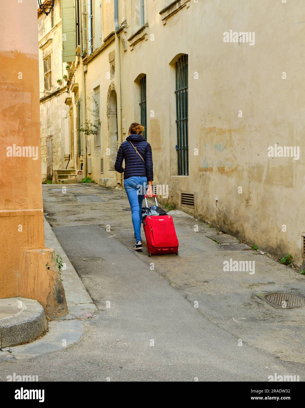 Jeune femme, marchant, tirant une valise roulante rouge dans l'étroite rue de la vieille ville sur Arles, France, Banque D'Images