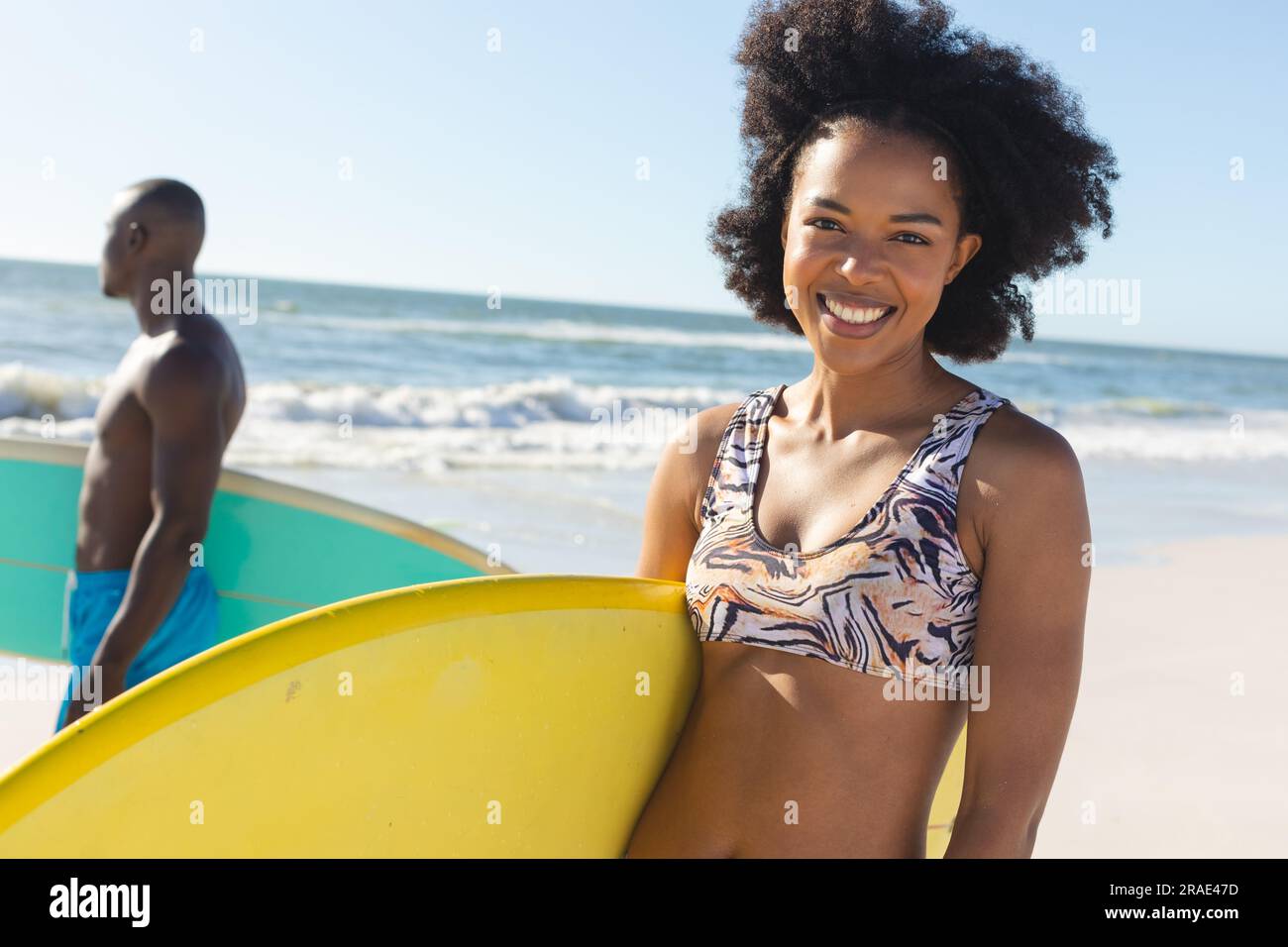 Portrait de femme afro-américaine heureuse tenant la planche de surf debout sur la plage ensoleillée avec surfeur masculin Banque D'Images