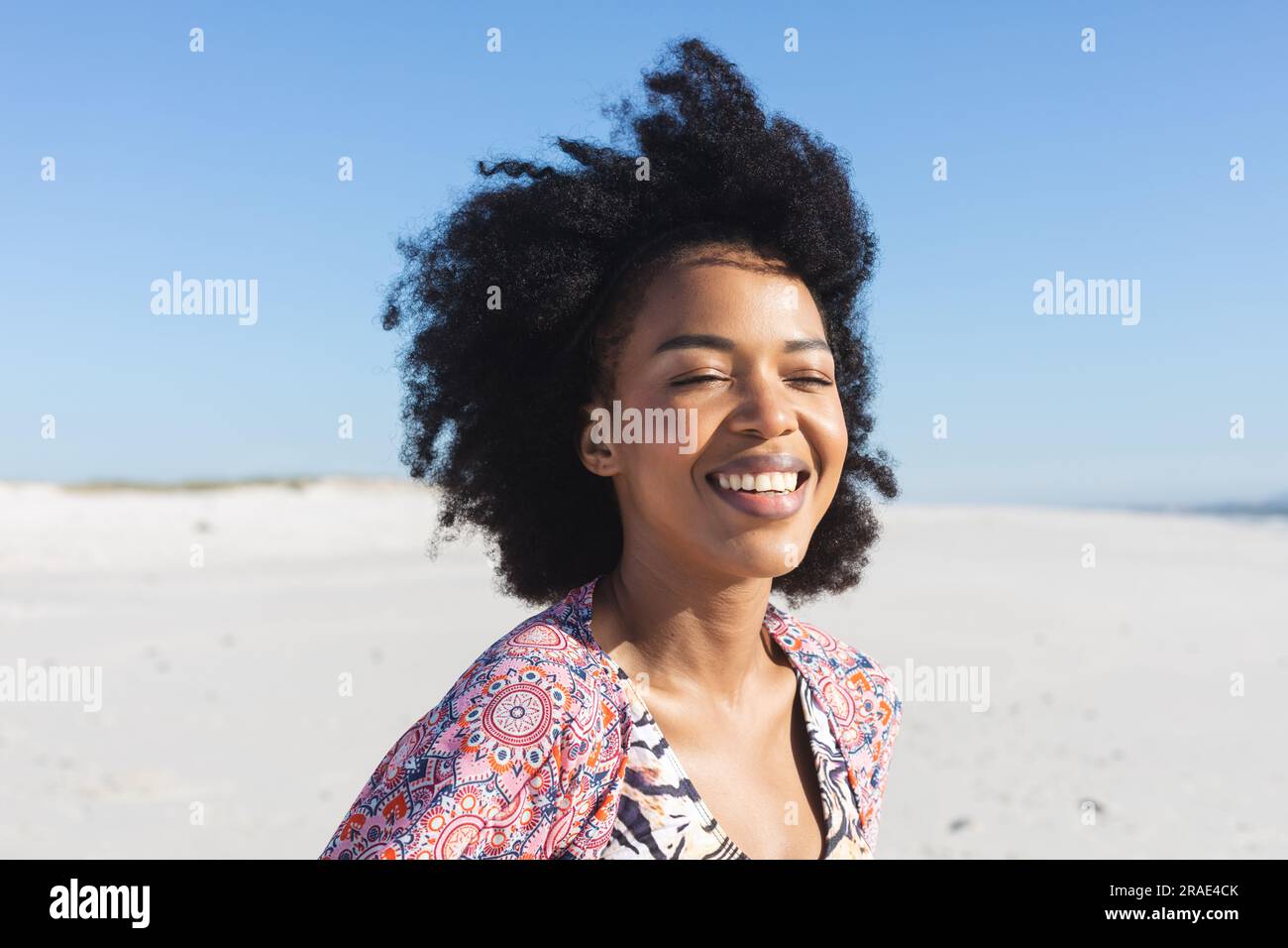 Heureuse femme afro-américaine assise sur une plage ensoleillée avec les yeux fermés, souriant Banque D'Images