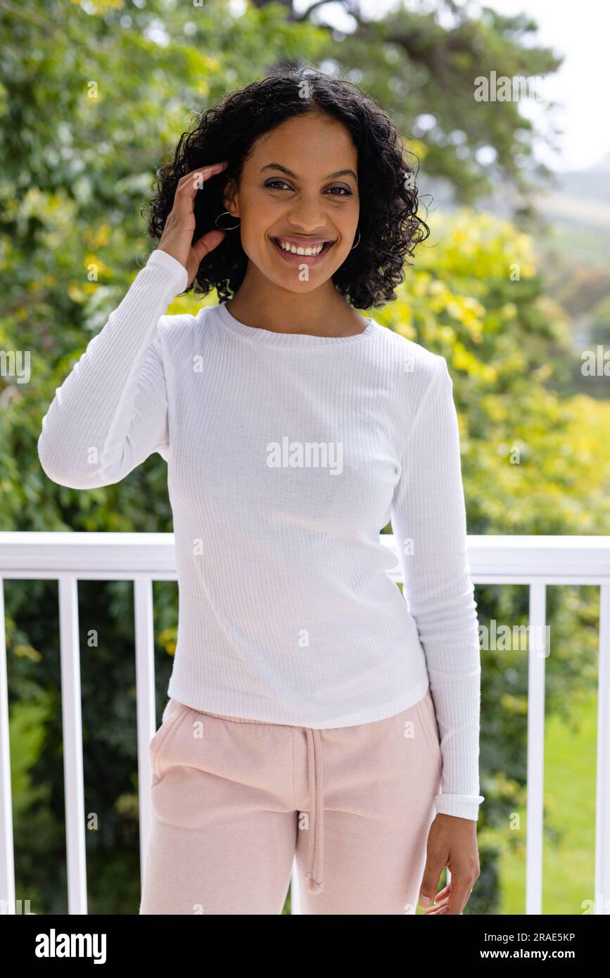 Portrait de souriante femme biraciale debout sur le balcon ensoleillé à la maison devant la cime des arbres Banque D'Images