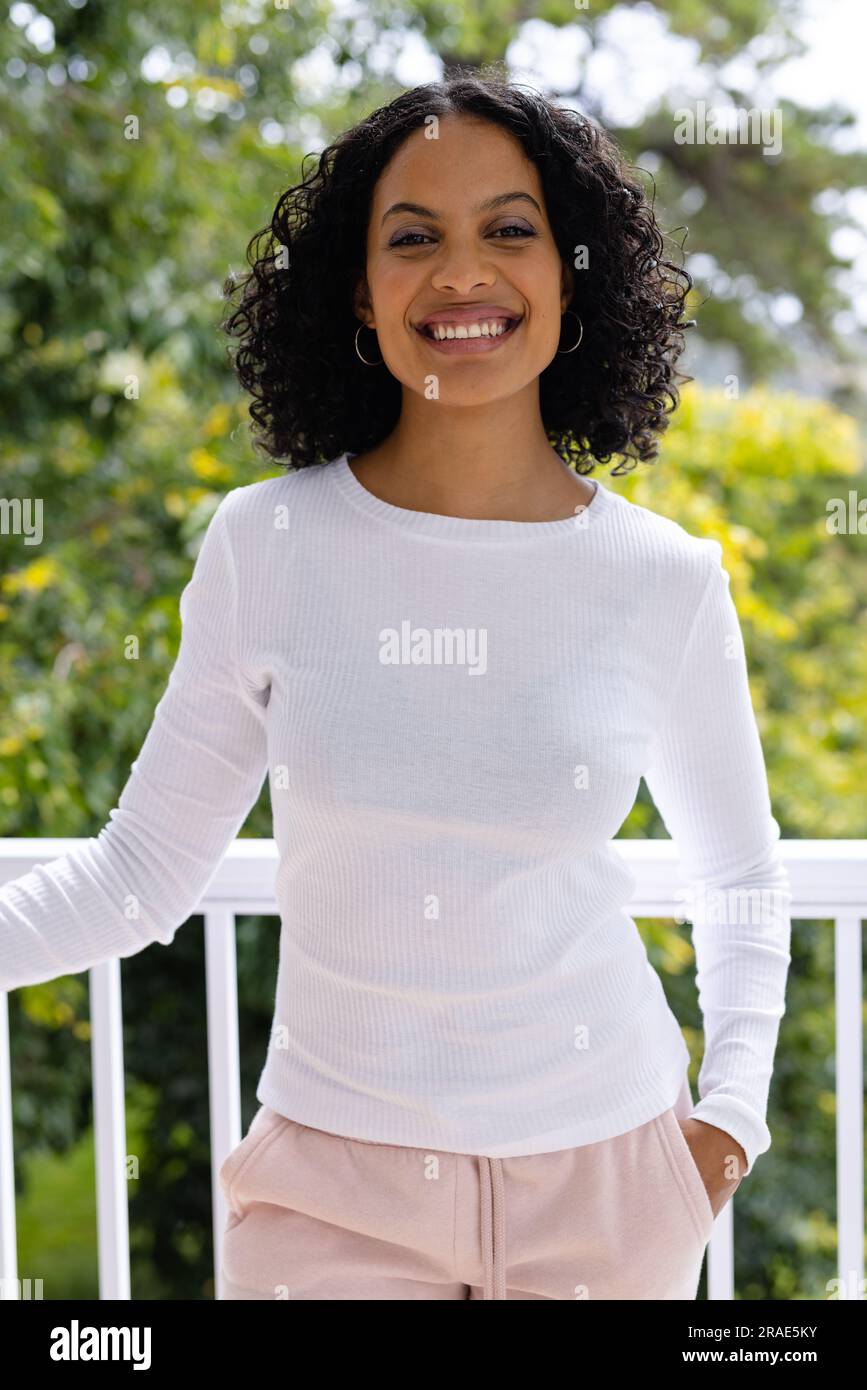 Portrait de souriante femme biraciale debout sur le balcon ensoleillé à la maison devant la cime des arbres Banque D'Images