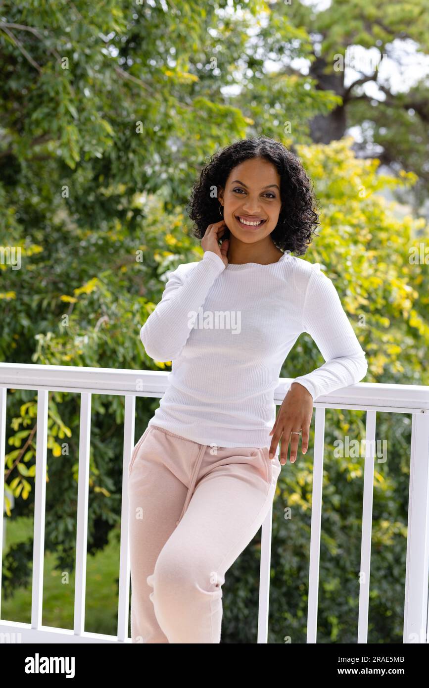 Heureuse femme biracial dans des pantoufles se relaxant sur le balcon à la maison avec cime des arbres en arrière-plan Banque D'Images