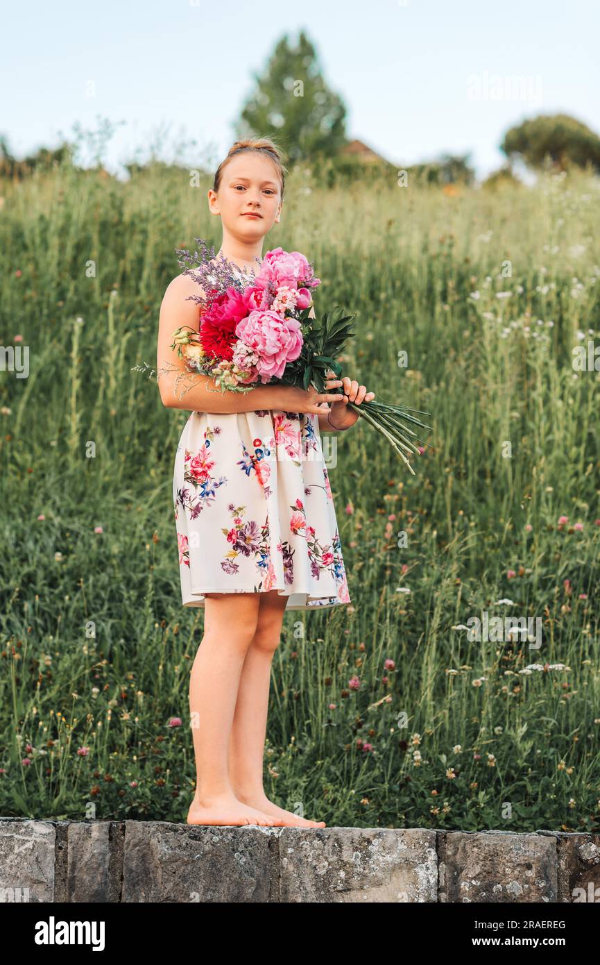 Portrait d'été d'adorable petite fille portant une belle robe occasionnelle, tenant un grand bouquet de pivoines, posant à l'extérieur Banque D'Images