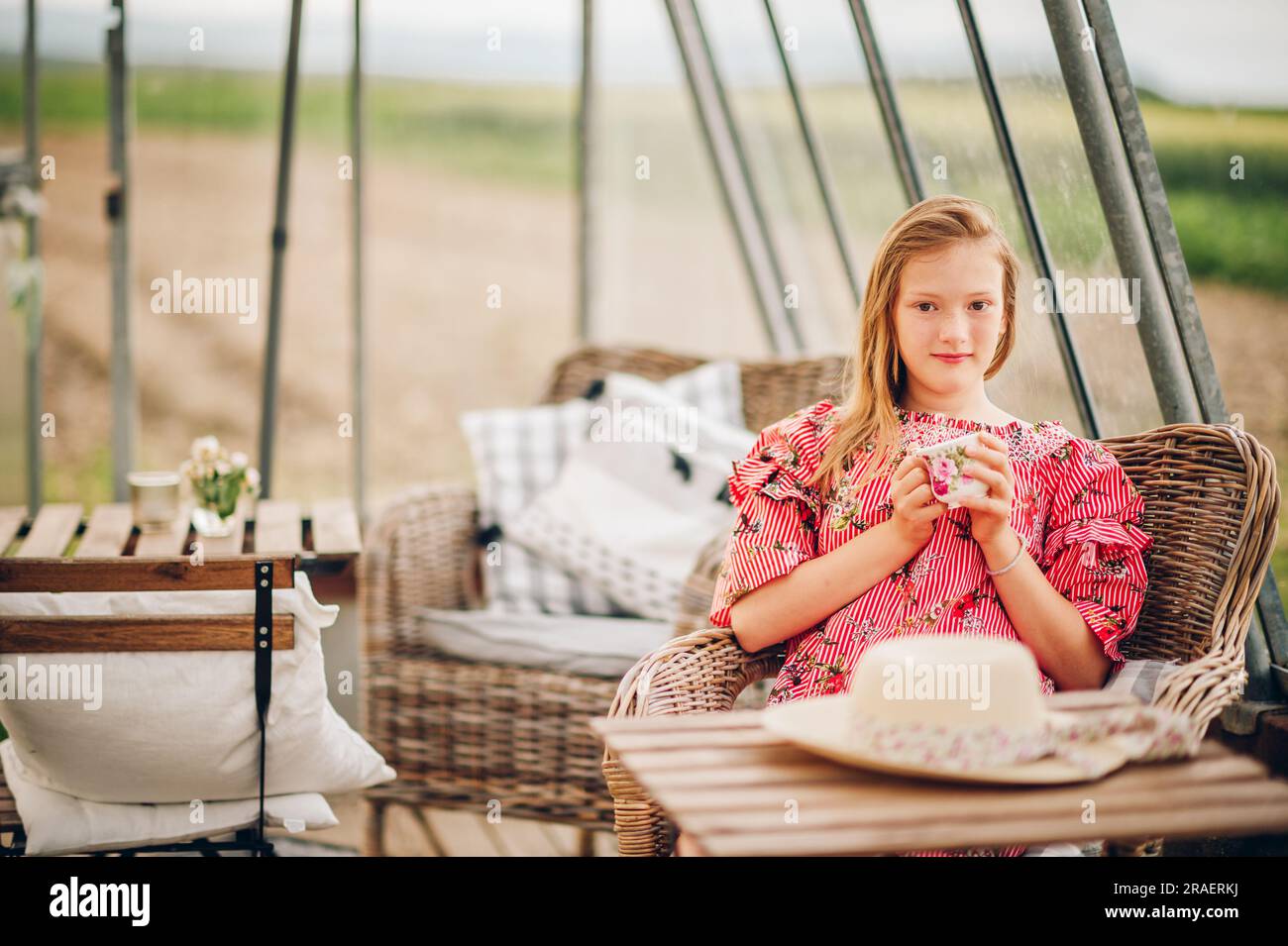 Jolie petite fille avec une tasse de thé reposant sur la terrasse d'été, portant une robe à rayures rouges, image rustique de style Banque D'Images