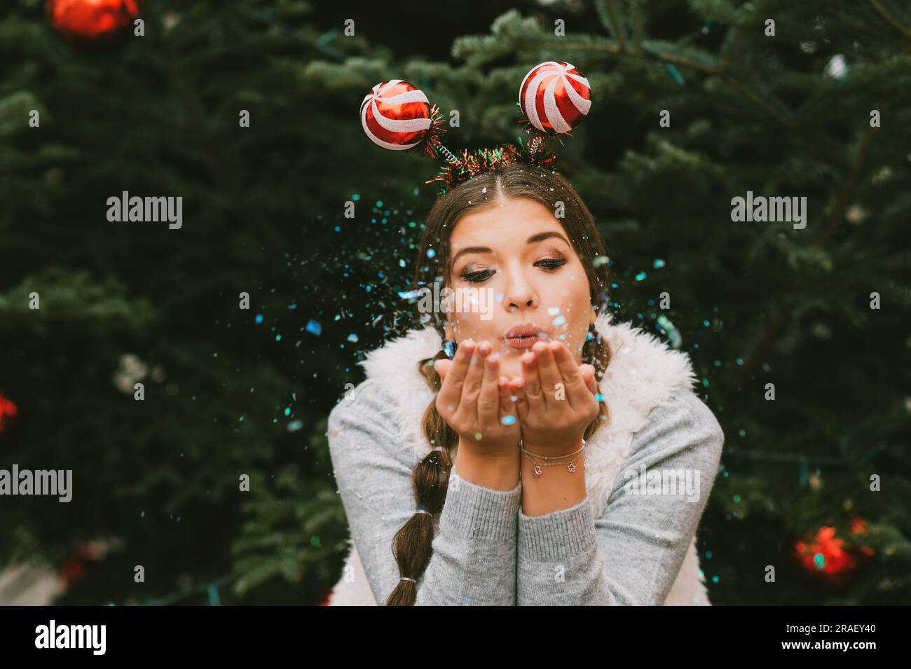 Portrait de fête de la jeune femme heureuse posant à l'extérieur par l'arbre de Noël, soufflant des paillettes, portant drôle de serre-tête de Noël Banque D'Images