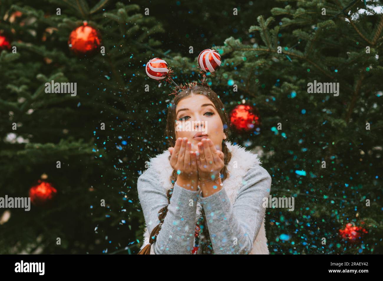 Portrait de fête de la jeune femme heureuse posant à l'extérieur par l'arbre de Noël, soufflant des paillettes, portant drôle de serre-tête de Noël Banque D'Images
