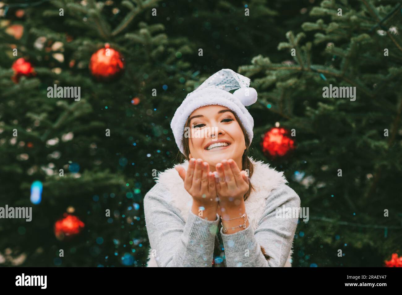 Portrait de fête de la jeune femme heureuse posant à l'extérieur près de l'arbre de Noël, soufflant des paillettes, portant le chapeau de père Noël argenté Banque D'Images