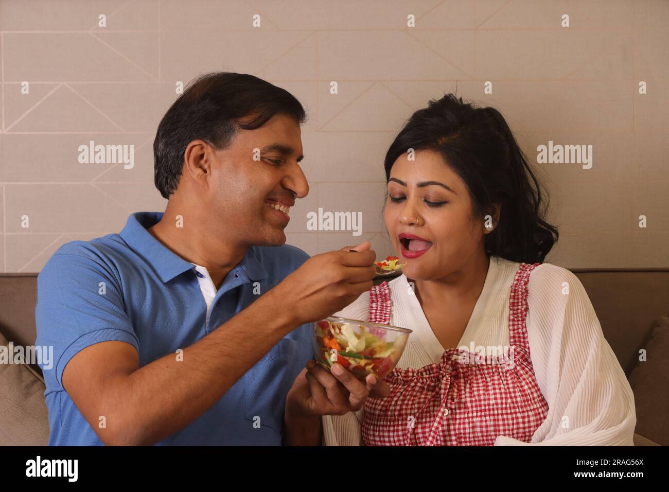 Heureuse femme de mari consciente de la santé indienne assise ensemble ayant un bol de salade verte assis sur le canapé dans un salon Banque D'Images