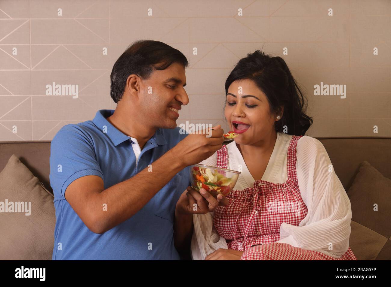 Heureuse femme de mari consciente de la santé indienne assise ensemble ayant un bol de salade verte assis sur le canapé dans un salon Banque D'Images