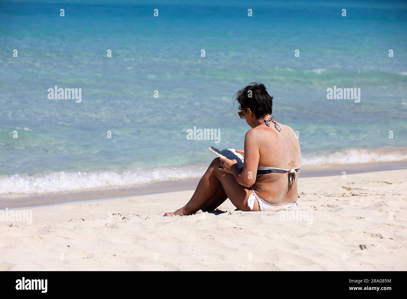 Femme en maillot de bain blanc assise sur le sable, bain de soleil et lecture d'un livre papier sur fond de vagues de mer. Loisirs sur la plage sur la côte tropicale Banque D'Images