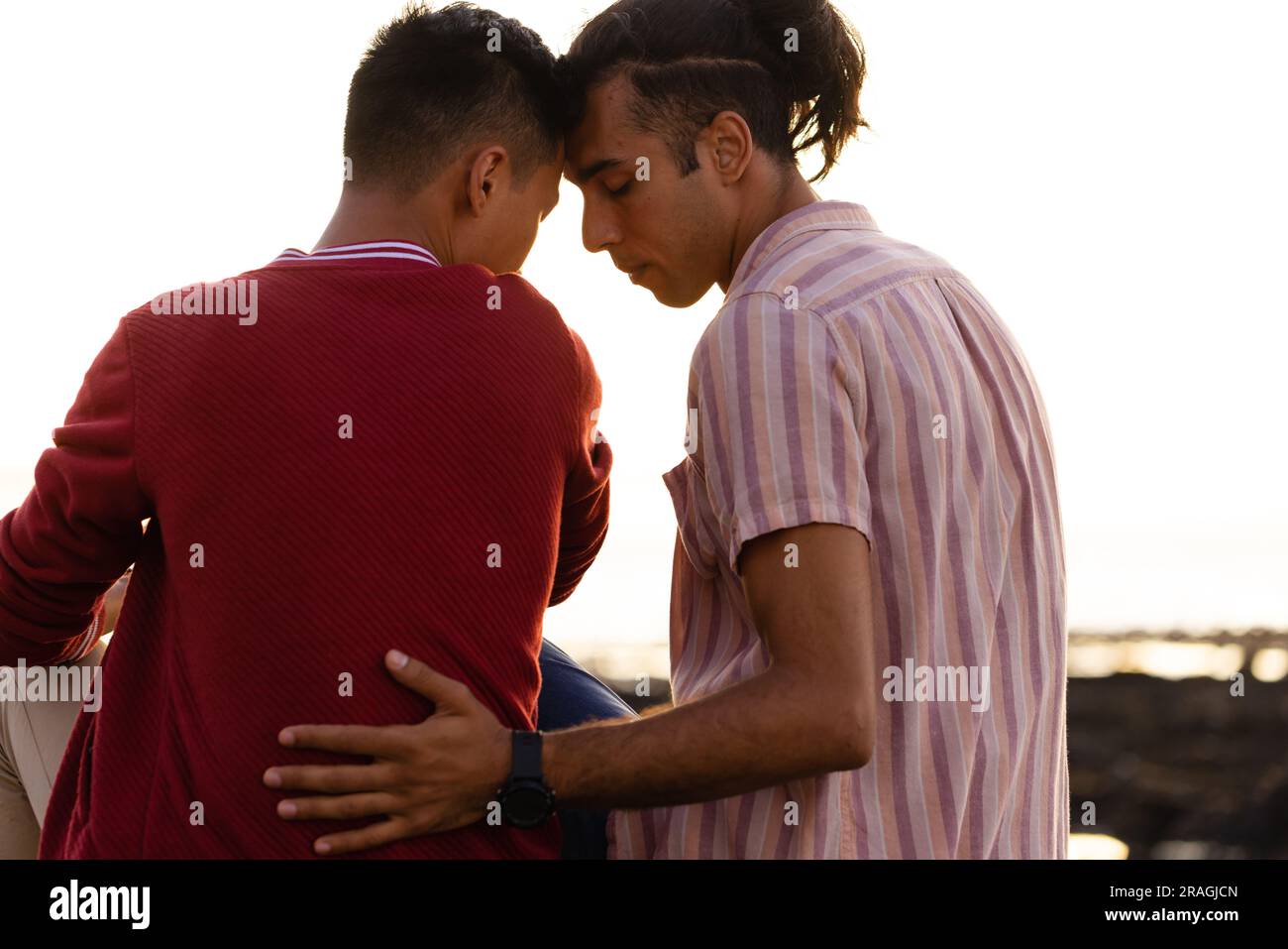 Joyeux couple masculin gay biracial assis sur des rochers et embrassant sur la plage au coucher du soleil Banque D'Images