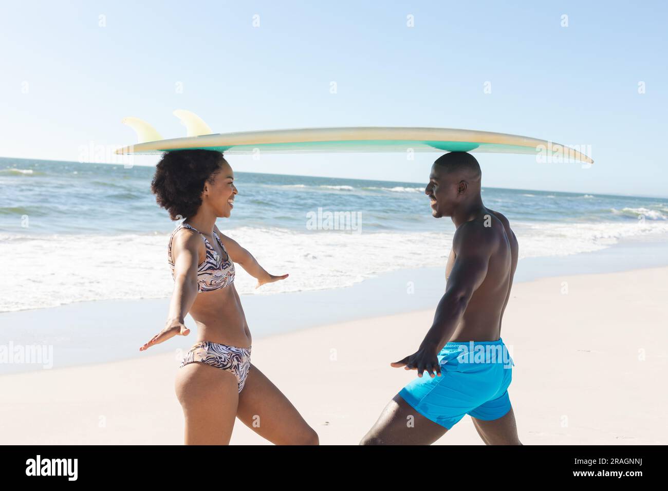 Heureux couple afro-américain équilibrant planche de surf sur les têtes, souriant sur la plage ensoleillée Banque D'Images