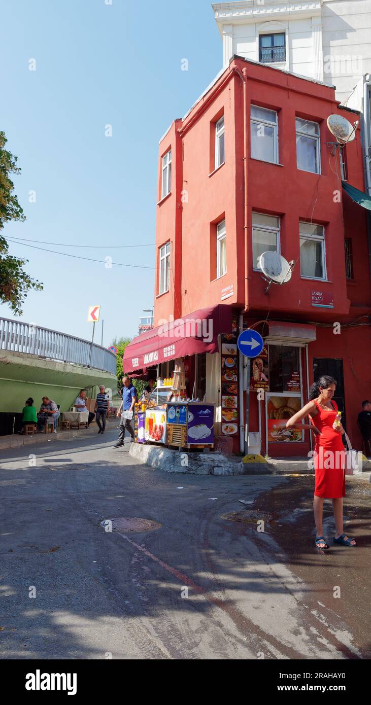 Femme en rouge debout à côté d'un bâtiment rouge abritant un kebab et un magasin de crème glacée, avec des gens buvant du thé turc sous une route, Istanbul, Turquie Banque D'Images