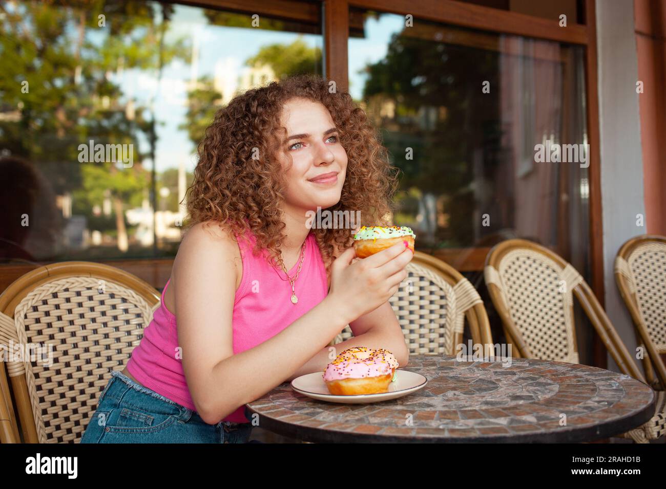 Portrait de belle jeune femme frisée dans le café sur la terrasse d'été, délicieux beignets colorés, pâtisseries sucrées. Pause déjeuner, rêveuse et heureuse. Thoughtfu Banque D'Images
