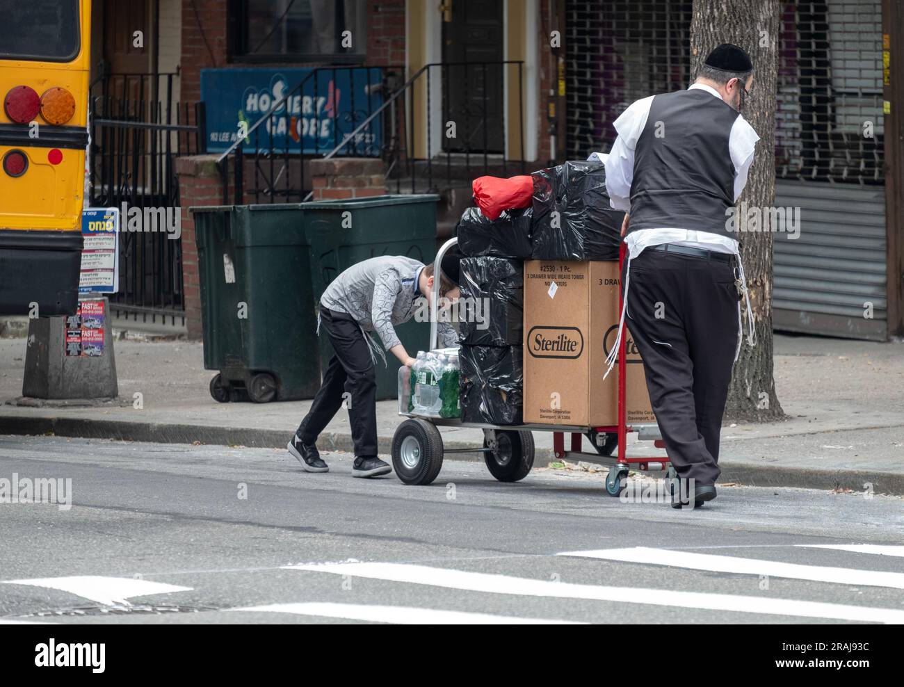 Un jeune garçon hassidique aide son père à déplacer des boîtes et des sacs d'un bus à leur domicile. Dans une rue animée de Brooklyn, New York. Banque D'Images