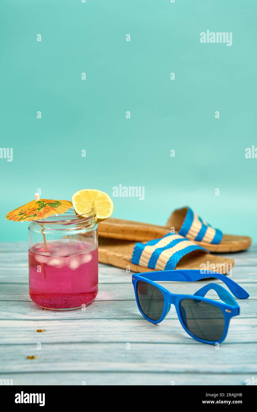 Boisson rafraîchissante de couleur rose avec un parapluie et une tranche de citron, lunettes de soleil bleues et sandales de plage, sur une table en bois et un fond vert. Banque D'Images