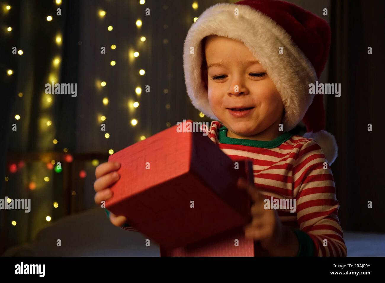 Un garçon de Noël en chapeau de père Noël souriant à la maison le soir. Nouvel an et jours fériés. Noël bonheur. Le garçon avec boîte cadeau s'amuser, profiter de Noël. Banque D'Images