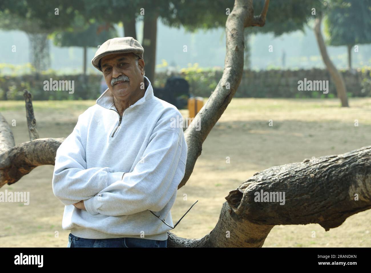 Vieil homme gai indien en bonne santé dans le parc debout à côté de l'arbre ayant du bon temps. Il porte une casquette de golf debout à côté d'une branche d'arbre. Banque D'Images