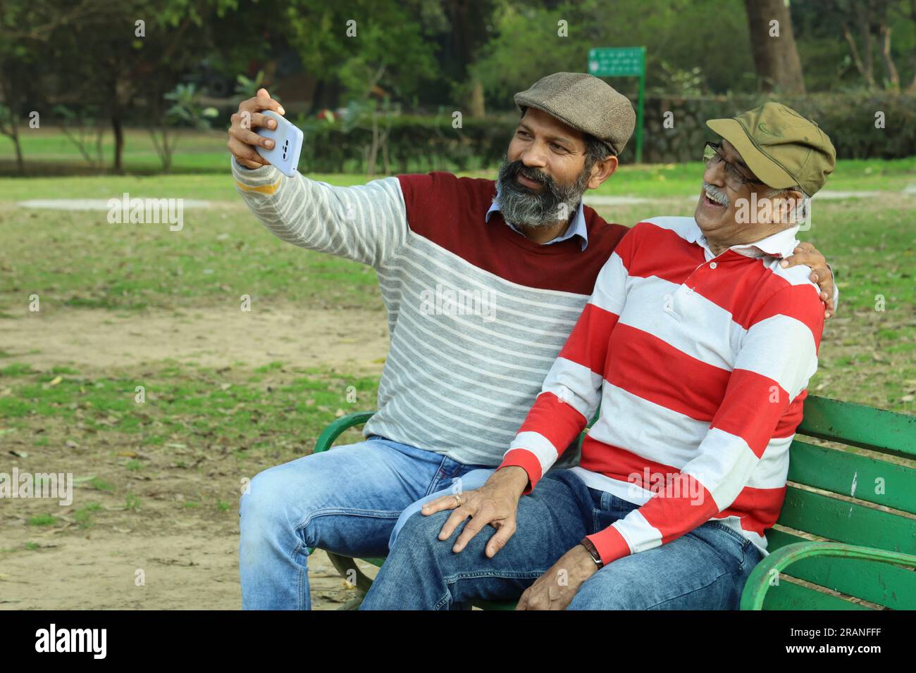 Vieux père et fils d'âge moyen assis sur un banc de parc tenant le téléphone portable en main. Appel vidéo et prise de selfie. Ils sont dans des verts luxuriants et la sérénité. Banque D'Images