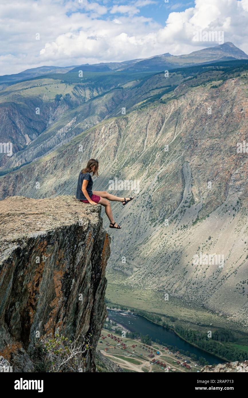 Photos dangereuses dans la nature. Une femme est assise sur le bord d'une falaise abrupte au-dessus d'une haute falaise et pend ses jambes. risque mortel pour la photographie. Summer Alta Banque D'Images