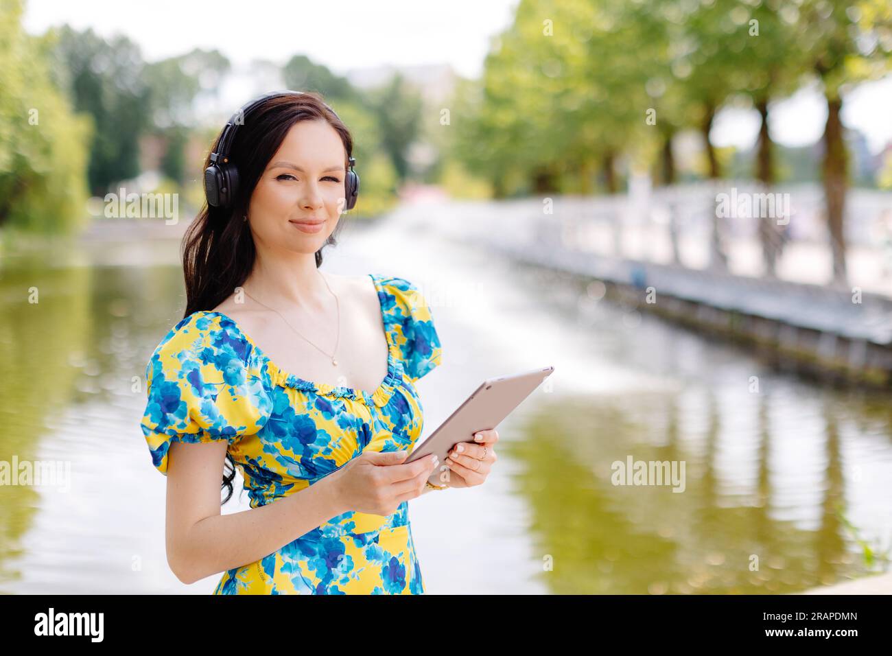 Jolie jeune femme avec des écouteurs écoutant de la musique via tablette numérique une journée ensoleillée près du lac. Banque D'Images
