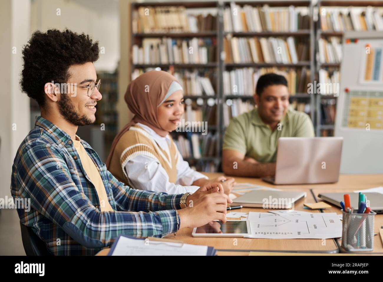 Étudiants migrants étudiant une langue étrangère assis ensemble à table avec des ordinateurs et des manuels scolaires Banque D'Images