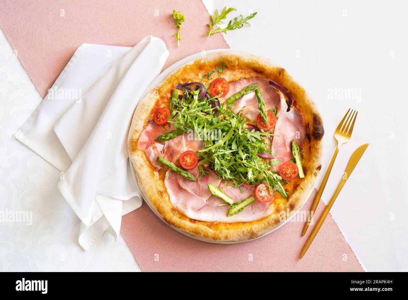 Vue de dessus d'une pizza au jambon avec jambon, asperges, tomates et roquette. Table de restaurant avec tissu rose et blanc, serviette blanche et couverts dorés. Banque D'Images