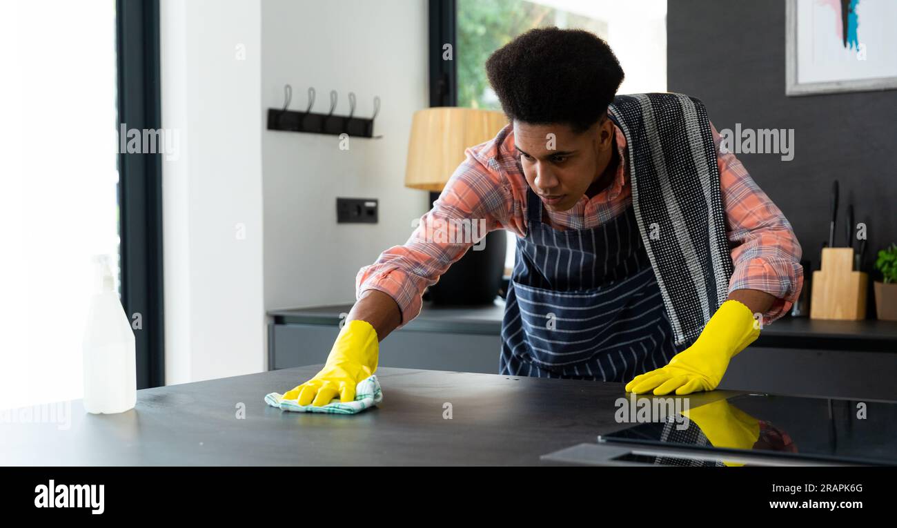 Homme biracial concentré portant un tablier et des gants en caoutchouc nettoyant le comptoir dans la cuisine ensoleillée. Nettoyage, hygiène, mode de vie sain et vie domestique, una Banque D'Images