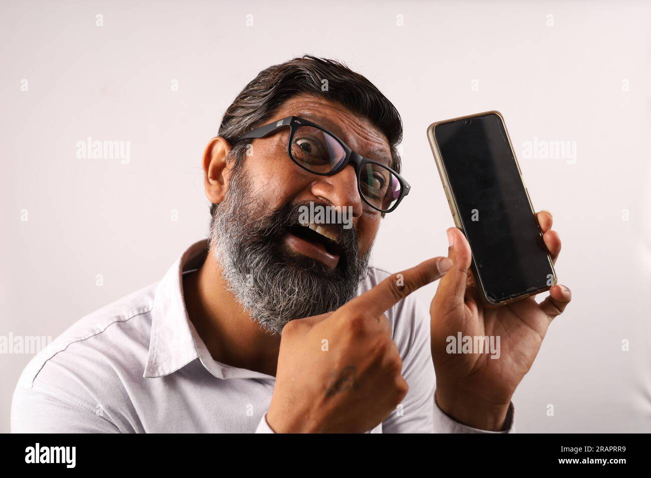 Homme barbu indien d'âge moyen portant une chemise tenant et montrant un téléphone portable dans sa main. Expressions funky sur un fond d'avion. Banque D'Images
