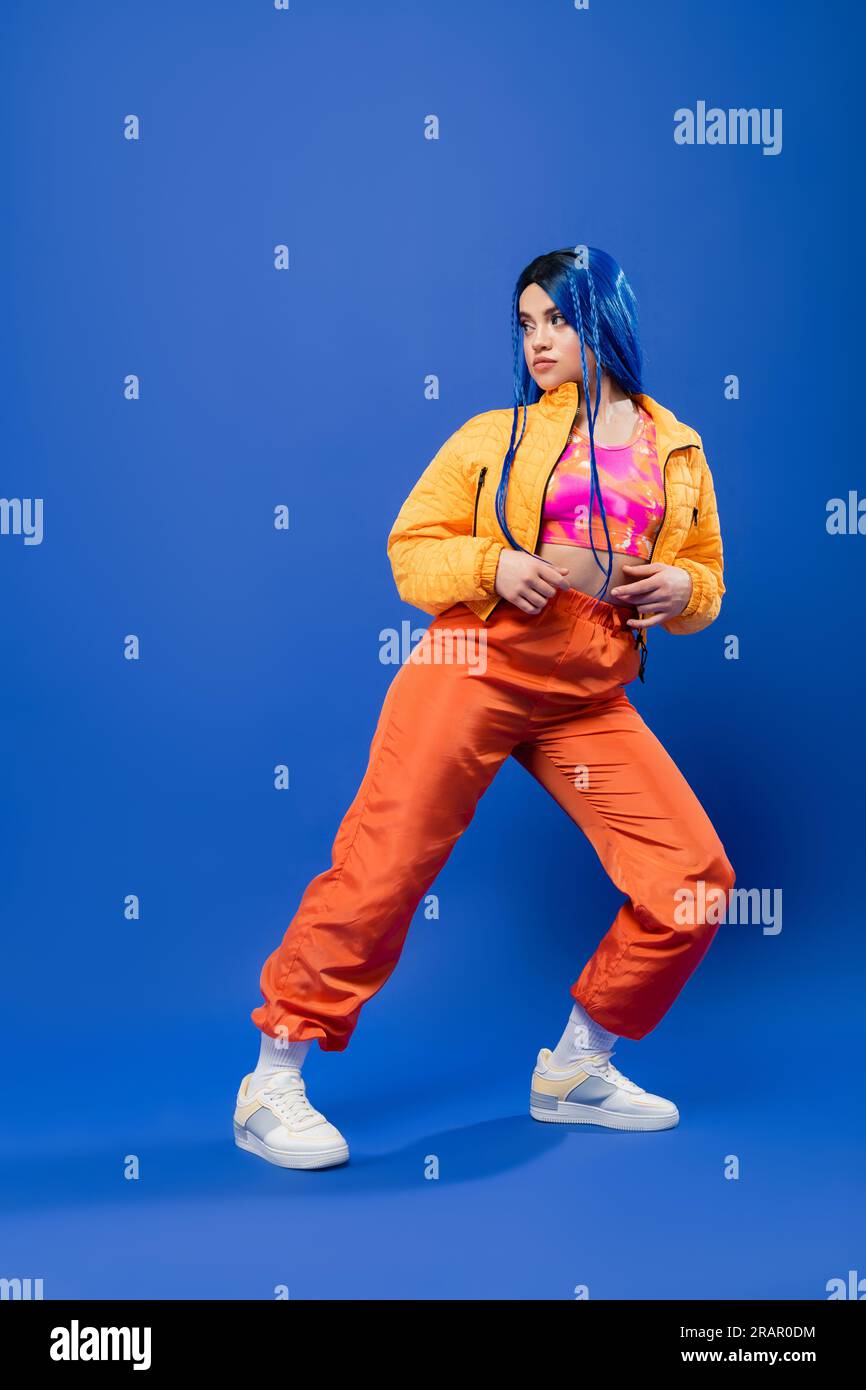 pleine longueur, cheveux teints, avant-gardiste de la mode, modèle féminin tatoué avec les cheveux bleus posant dans la veste bouffante et pantalon orange sur fond bleu, col vibrant Banque D'Images