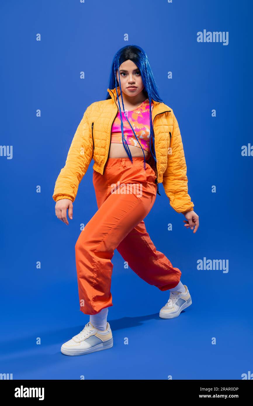 pleine longueur, avant-gardiste de la mode, jeune modèle féminin avec les cheveux bleus posant dans la veste bouffante et pantalon orange sur fond bleu, couleur vibrante, fash urbain Banque D'Images