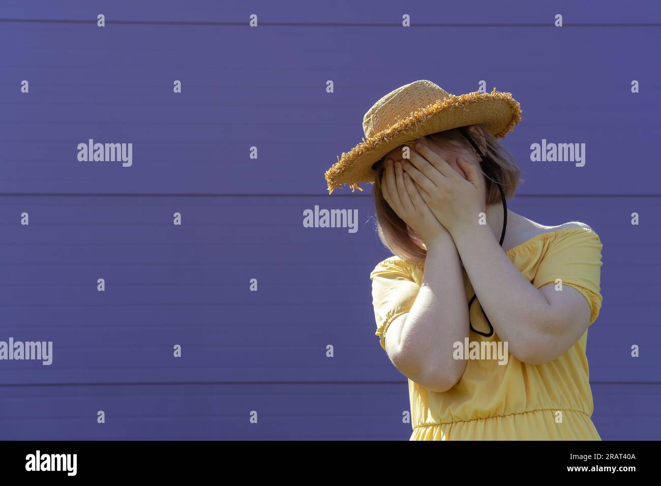 Une jeune fille asiatique frustrée dans un chapeau et une robe couvre son visage avec ses mains sur un fond violet. Concept de santé mentale. Banque D'Images