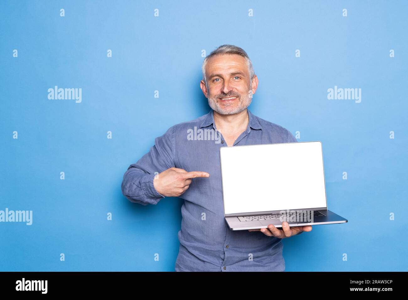 Joyeux homme de race blanche d'âge moyen montre le doigt sur l'ordinateur portable avec écran blanc, recommande l'ordinateur, isolé sur fond bleu.Quarantaine Covid-19 avec m Banque D'Images