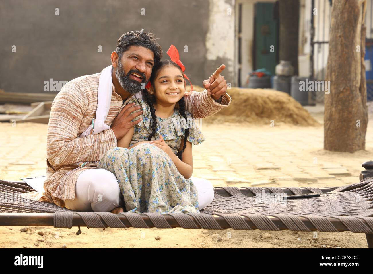 Heureuse famille indienne rurale dans le village. Père et fille assis ensemble souriant sur un lit bébé devant leur maison dans la cour avant. Père en pyjama kurta Banque D'Images