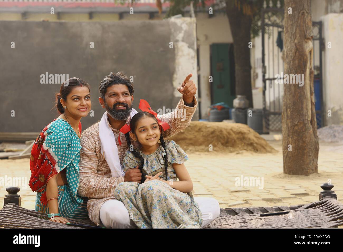 Heureuse famille indienne rurale assise ensemble à l'extérieur de leur chalet dans la journée souriant tout en regardant la caméra Banque D'Images