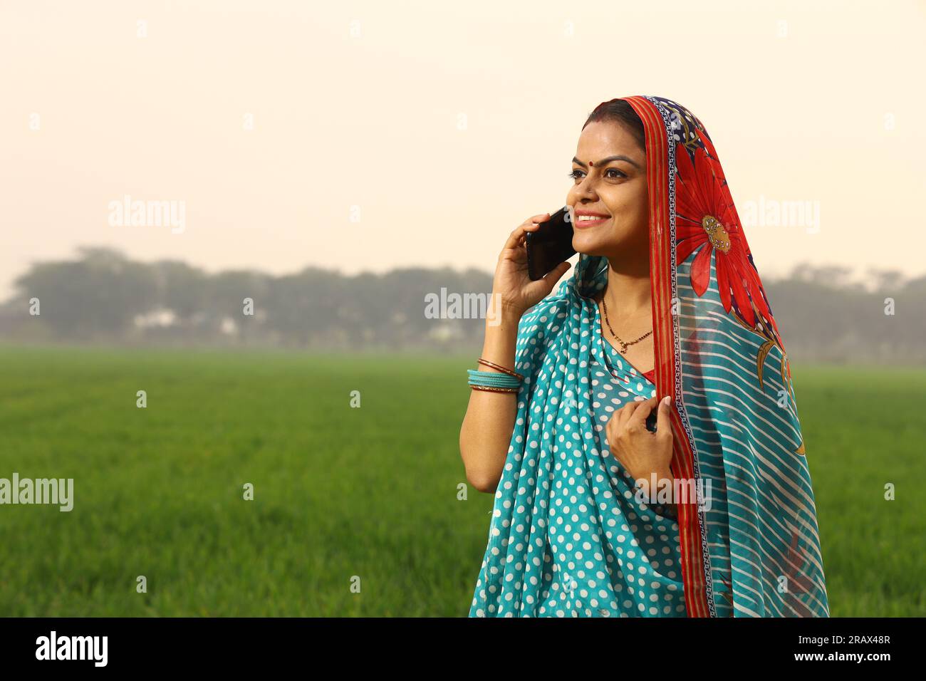 Heureuse femme indienne rurale debout dans un champ agricole de moutarde et surfant à travers le téléphone portable dans sa main. Banque D'Images