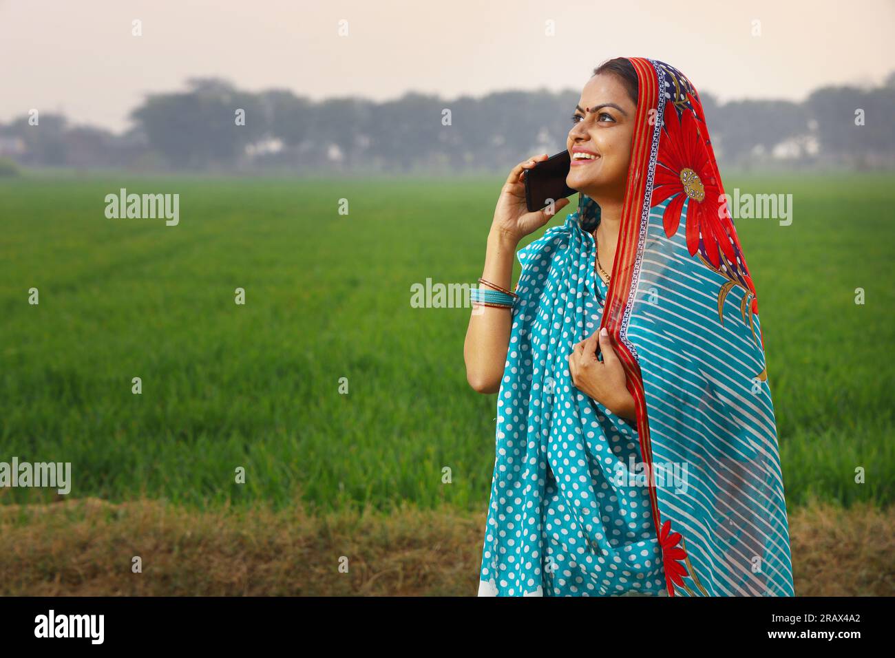 Heureuse femme indienne rurale debout dans un champ agricole de moutarde et surfant à travers le téléphone portable dans sa main. Banque D'Images