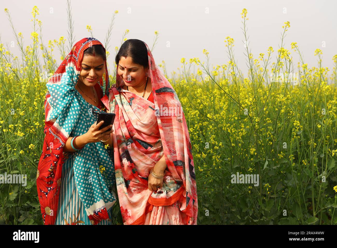 Femmes indiennes rurales heureuses debout dans un champ agricole de moutarde et surfant à travers le téléphone portable dans leur main. Banque D'Images