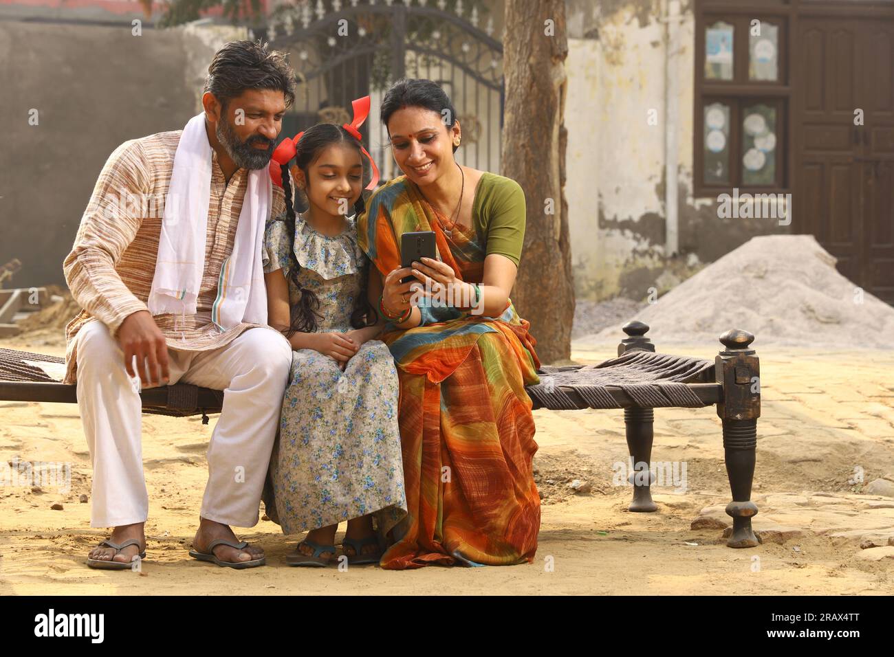 Heureux Indien famille rurale de mère, père et fille assis ensemble souriant sur lit à l'extérieur de leur maison dans la cour avant en utilisant le téléphone portable. Banque D'Images