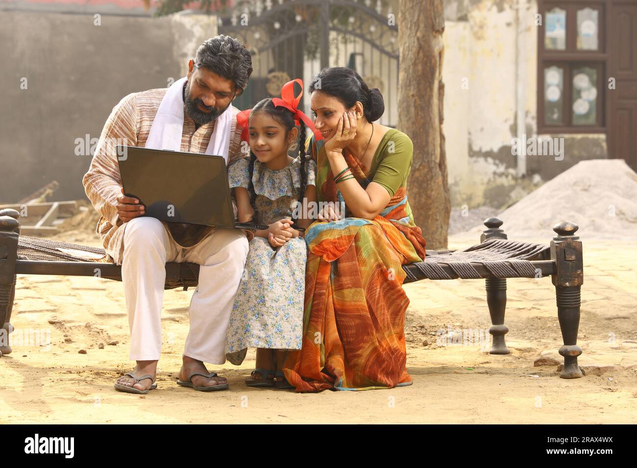 Heureux Indien famille rurale de mère, père et fille assis ensemble souriant sur lit à l'extérieur de leur maison dans la cour avant en utilisant un ordinateur portable. Banque D'Images