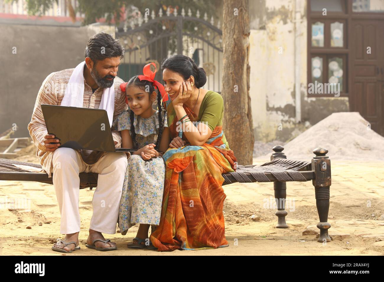 Heureux Indien famille rurale de mère, père et fille assis ensemble souriant sur lit à l'extérieur de leur maison dans la cour avant en utilisant un ordinateur portable. Banque D'Images